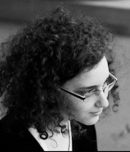 Chally, in black and white, gazes to the right of the camera. It's a slightly high angle shot. She is wearing glasses and a black dress and top, of which only a little is visible. The base of a pillar is just visible behind her.